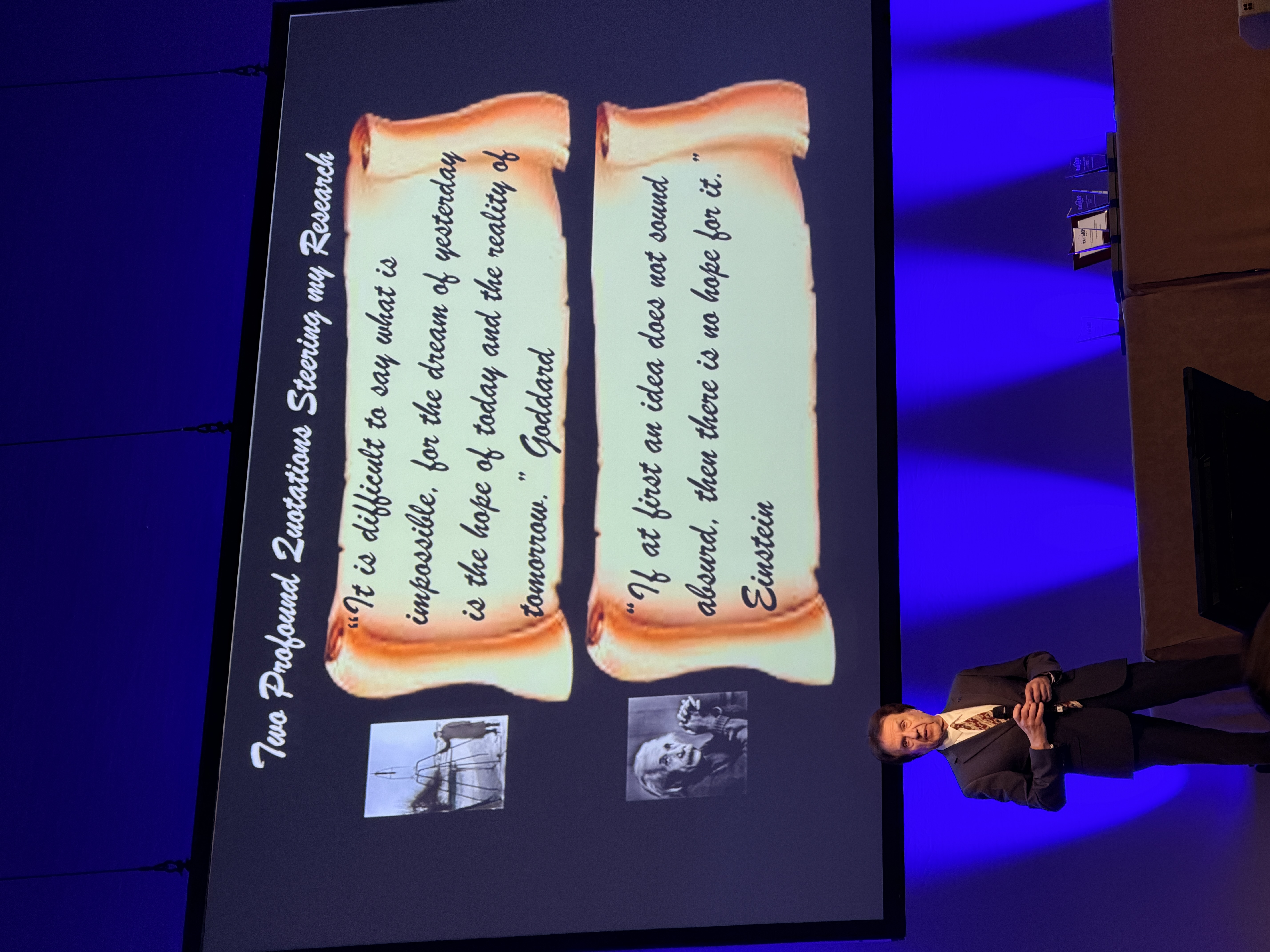 A speaker on a blue-lit stage beneath a slide containing research-themed quotations.